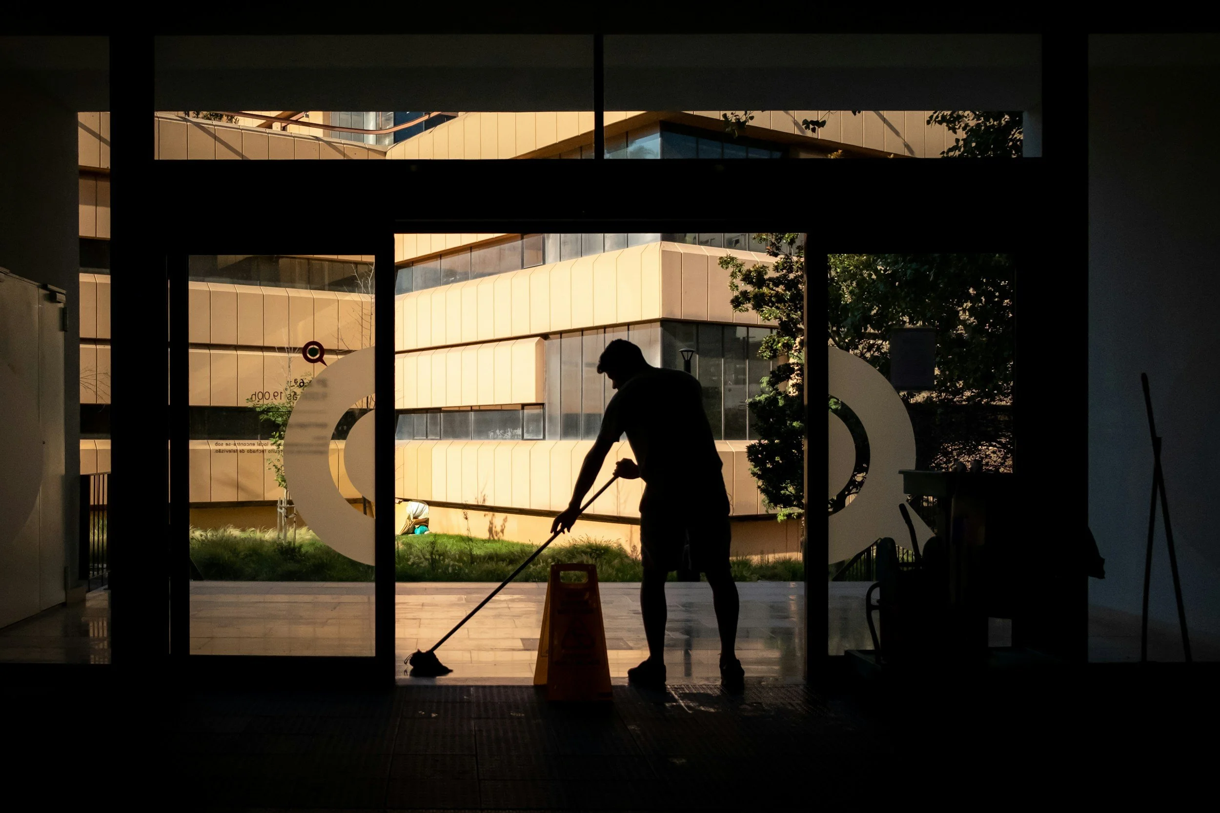 A silhouette of a janitor mopping the floor near a large glass door with a yellow building outside and some greenery.