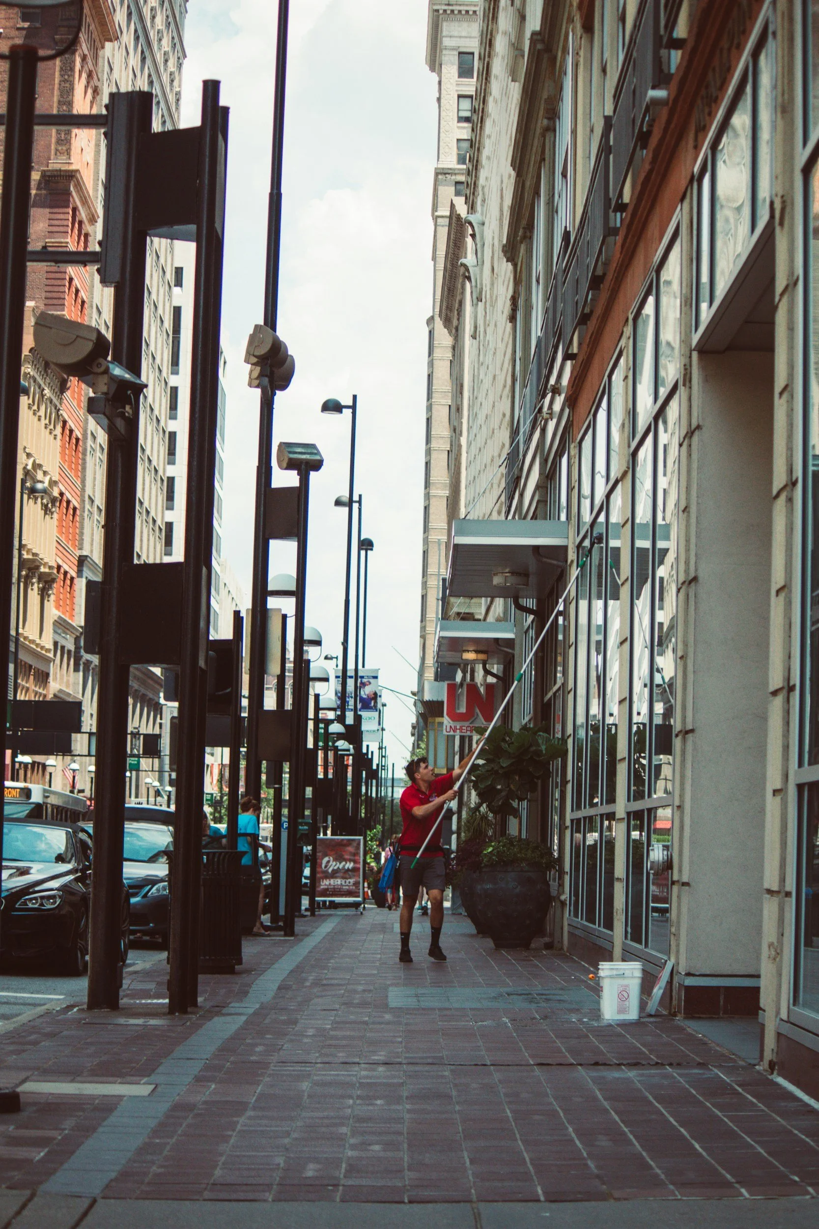 A worker cleaning the windows of a building with a long pole and a squeegee on a city sidewalk.