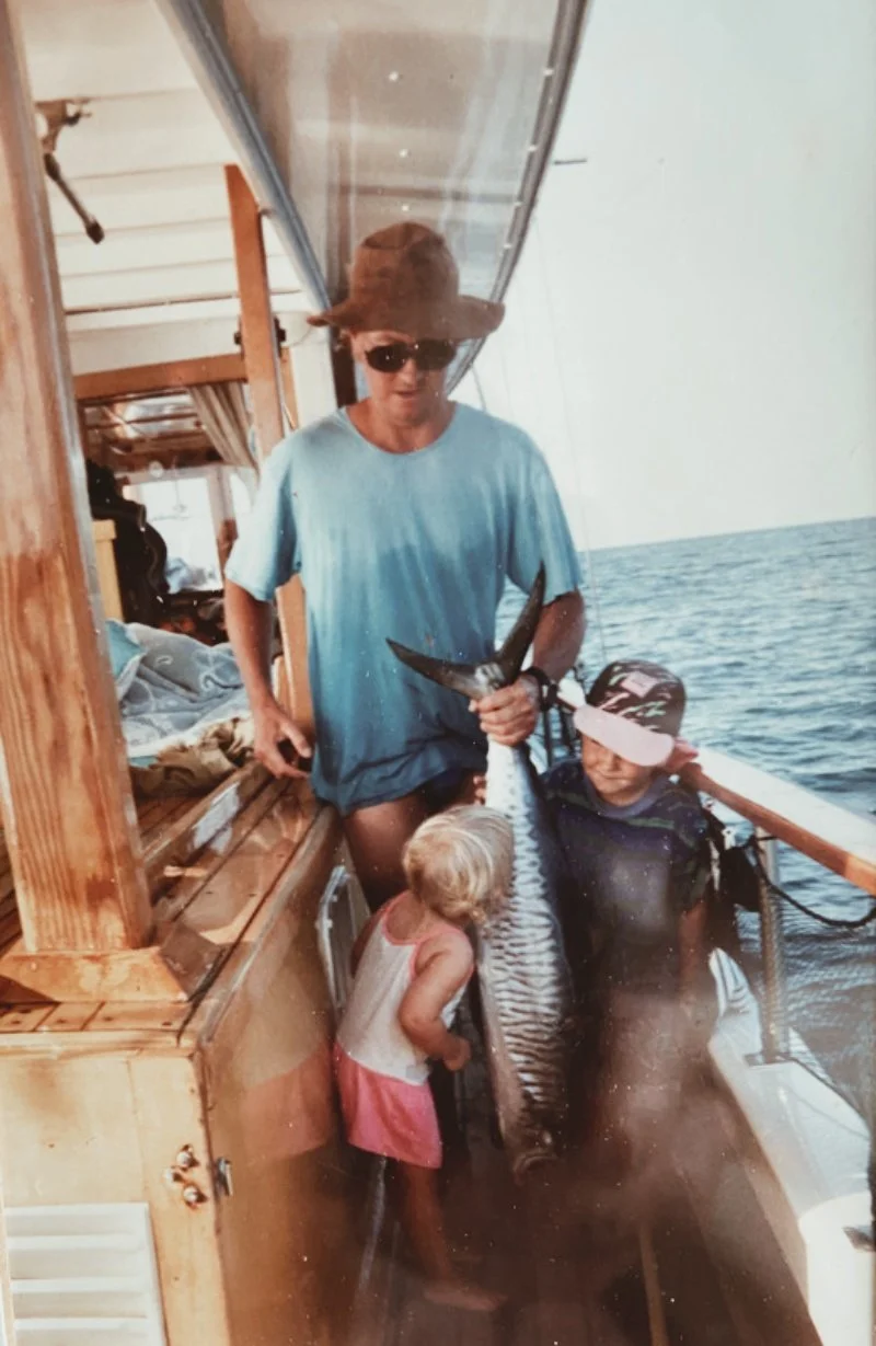 A man wearing sunglasses, a wide-brimmed hat, and a blue shirt holding a striped fish on a boat, with a young girl touching the fish and another girl wearing a cap nearby, over open water.