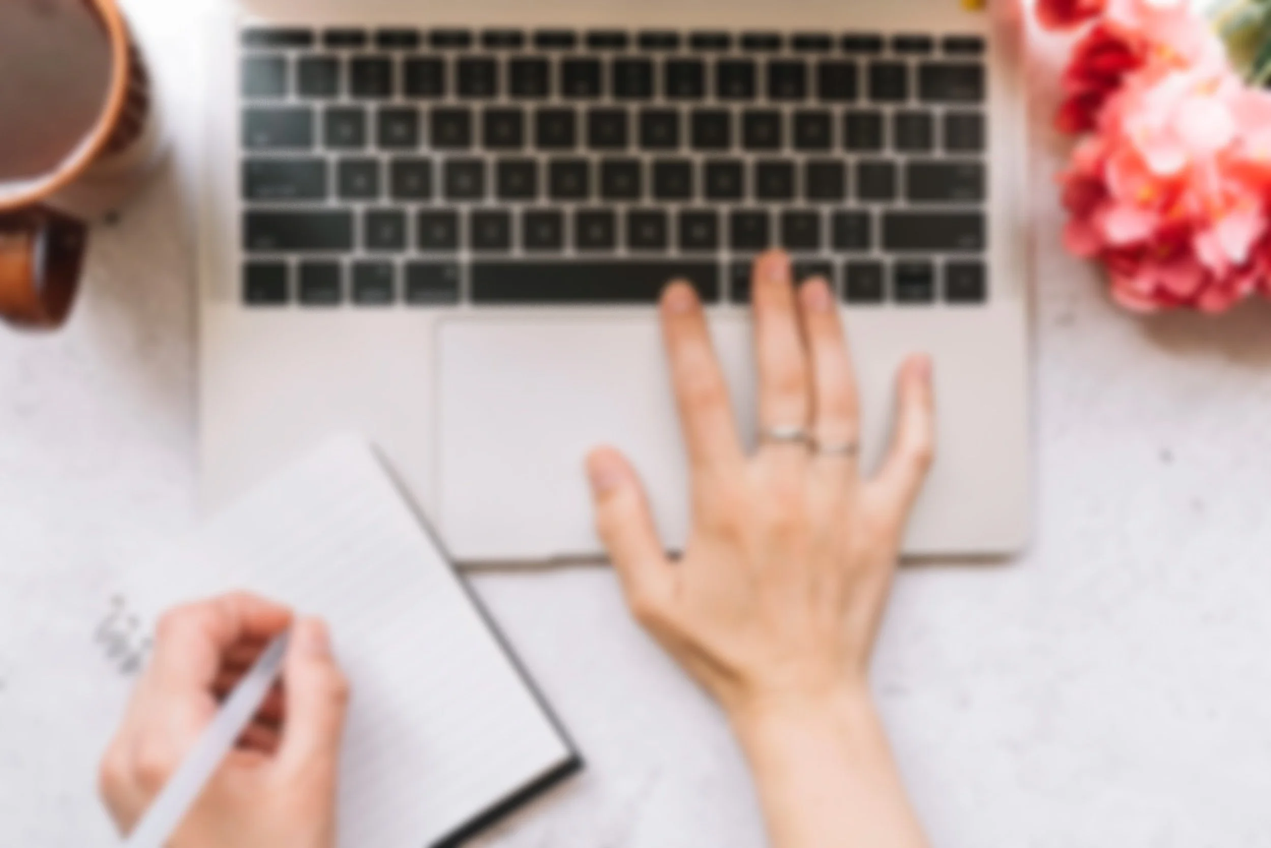 Blurred image of a person's hand typing on a laptop keyboard, with a notebook and pen in the opposite hand, a mug on the left, and pink flowers on the right.
