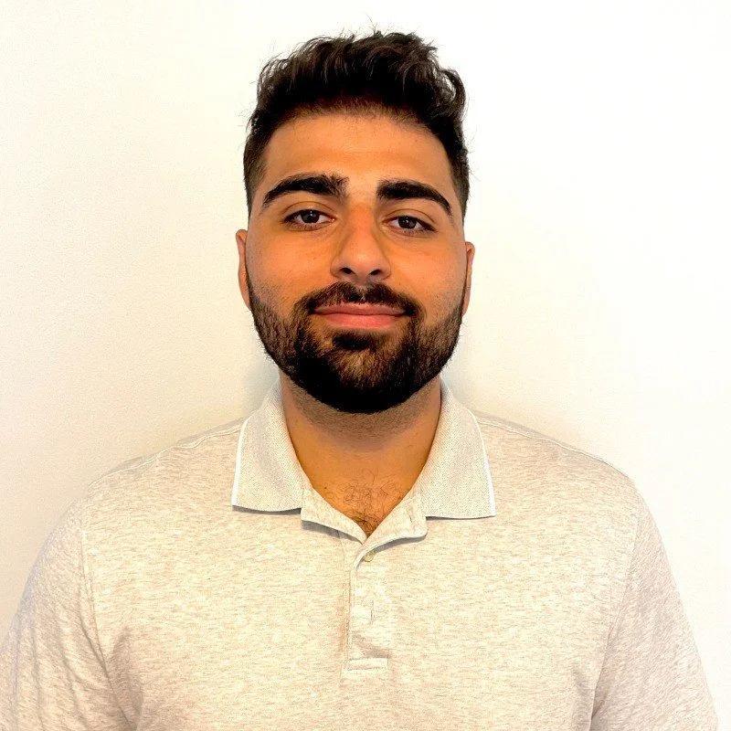 A young man with dark hair, a beard, and a mustache, smiling slightly, wearing a light-colored polo shirt standing against a plain white wall.
