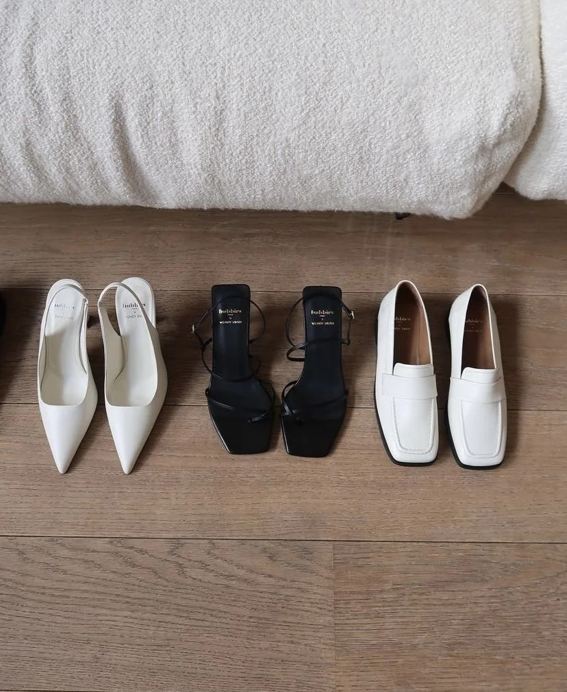 Four pairs of women's shoes arranged on a wooden floor in front of a bed: white pointed-toe heels, black strappy block heels, white loafers, and cream-colored loafers.