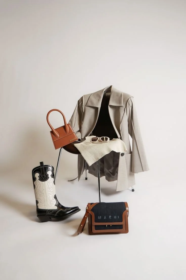 Fashion display with a beige coat, black top, white sunglasses, a beige sweater, and accessories including a small brown handbag, black and white cowboy boots, a black and brown Marni handbag, and a pair of cream-colored sunglasses on a white surface against a plain white background.
