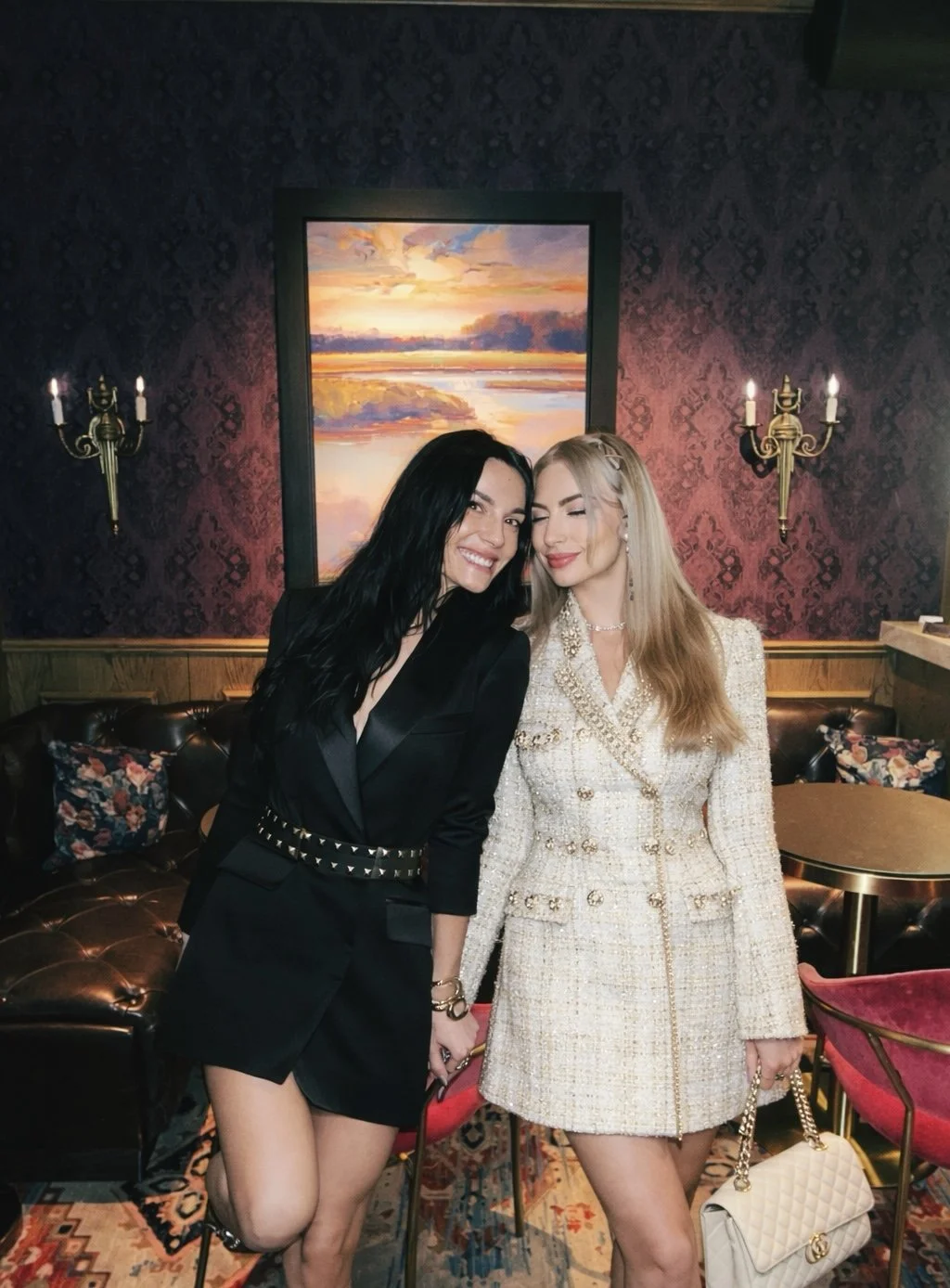 Two women dressed elegantly, smiling and holding hands inside a dimly lit, ornate room with patterned wallpaper, flower-patterned cushions, a painting of a colorful sunrise or sunset, and wall-mounted candle-style lights.