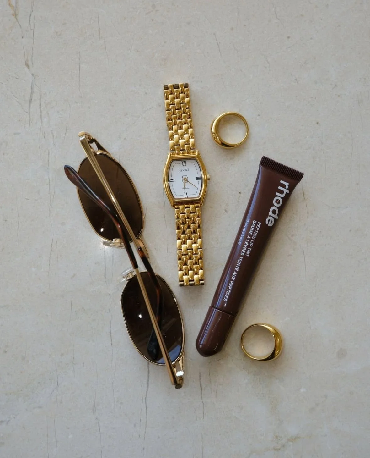 A collection of personal accessories including a pair of sunglasses, a gold wristwatch, a gold ring, a tube of brown lip balm, and another gold ring, all arranged on a light-colored surface.