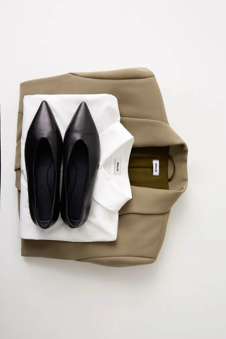 A pair of black pointed-toe flats, white t-shirt, beige blazer, and tan tote bag on a white background.