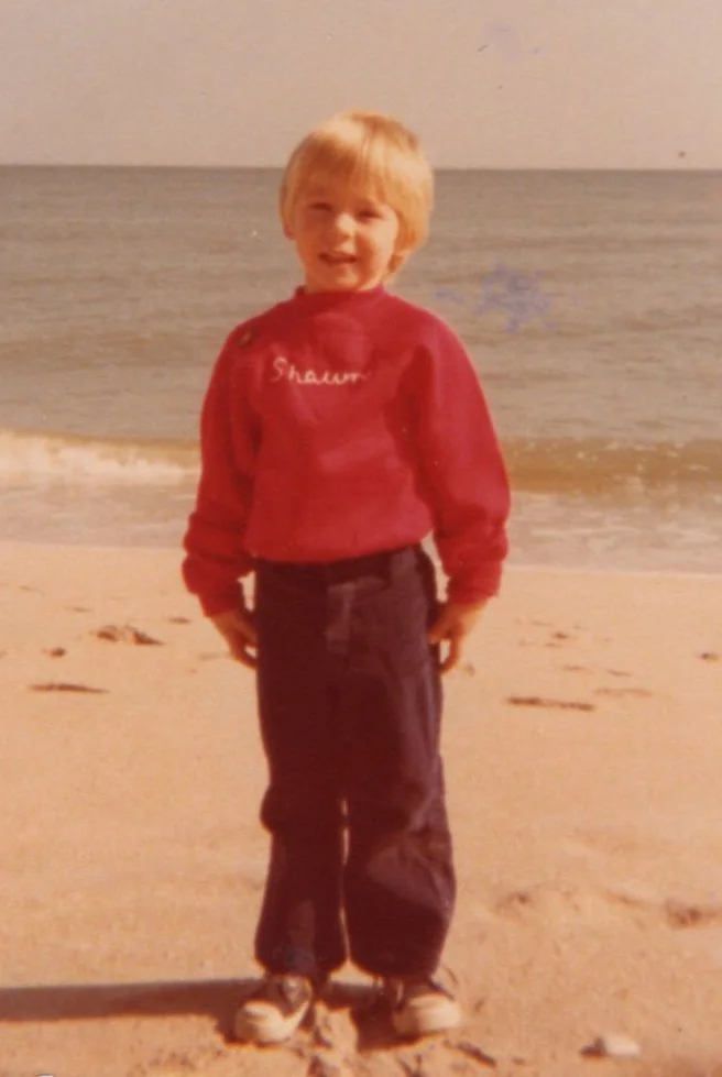 A young boy with blonde hair smiling on a sandy beach near the ocean during daytime, wearing a red sweatshirt with 'Shawn' embroidered on it and dark pants.