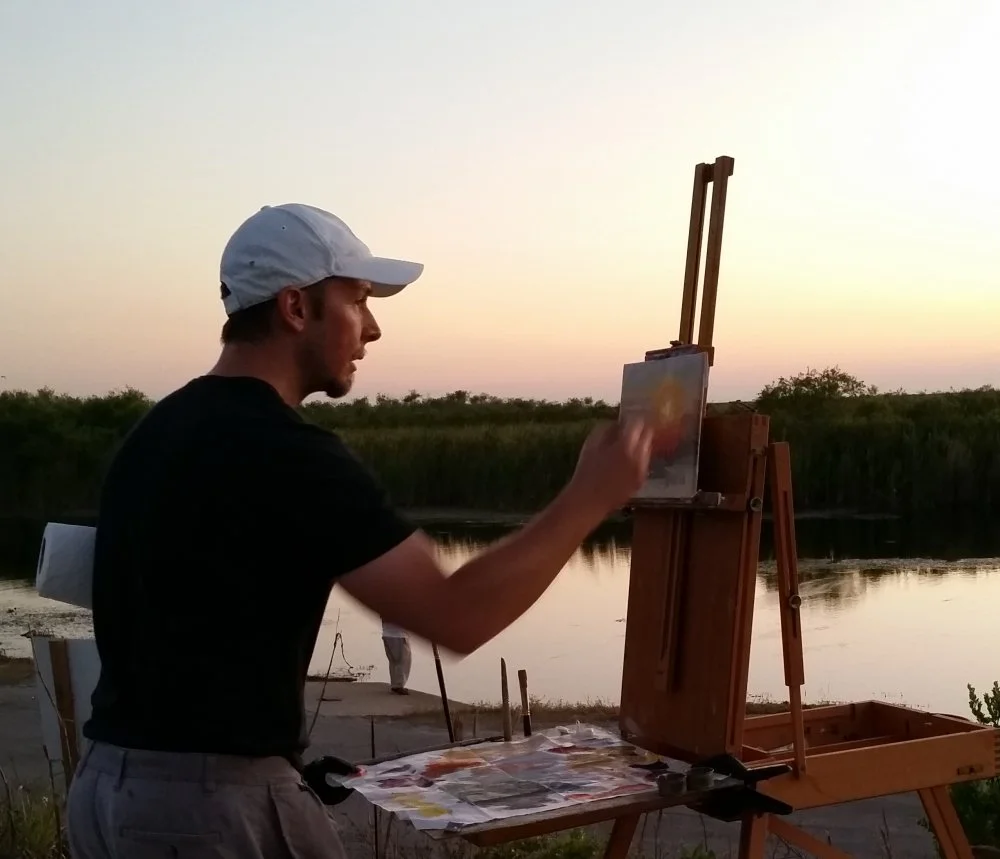 A man wearing a white cap and black shirt painting outdoors at sunset, standing at an easel with a landscape scene near a river with green trees in the background.