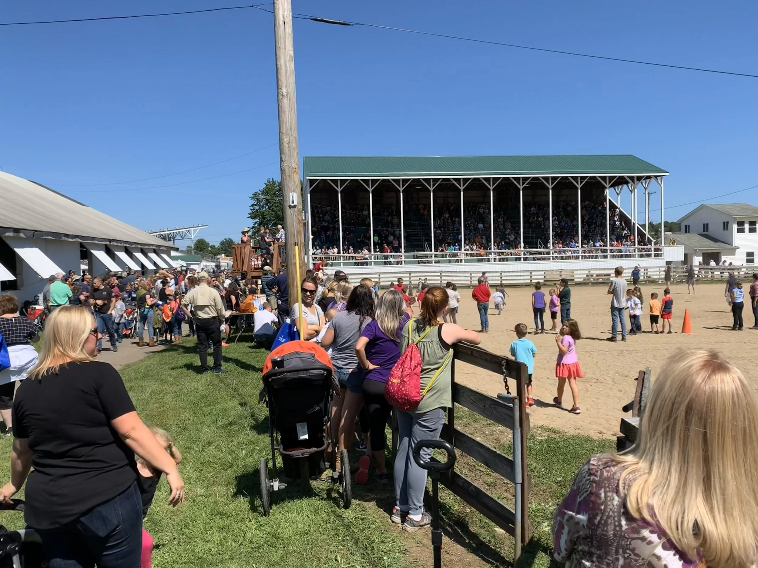 Crowd of people gathered at a rodeo arena on a sunny day, with some watching from the bleachers and others standing near the fence, children playing on the dirt field, and a few people talking near a pole.