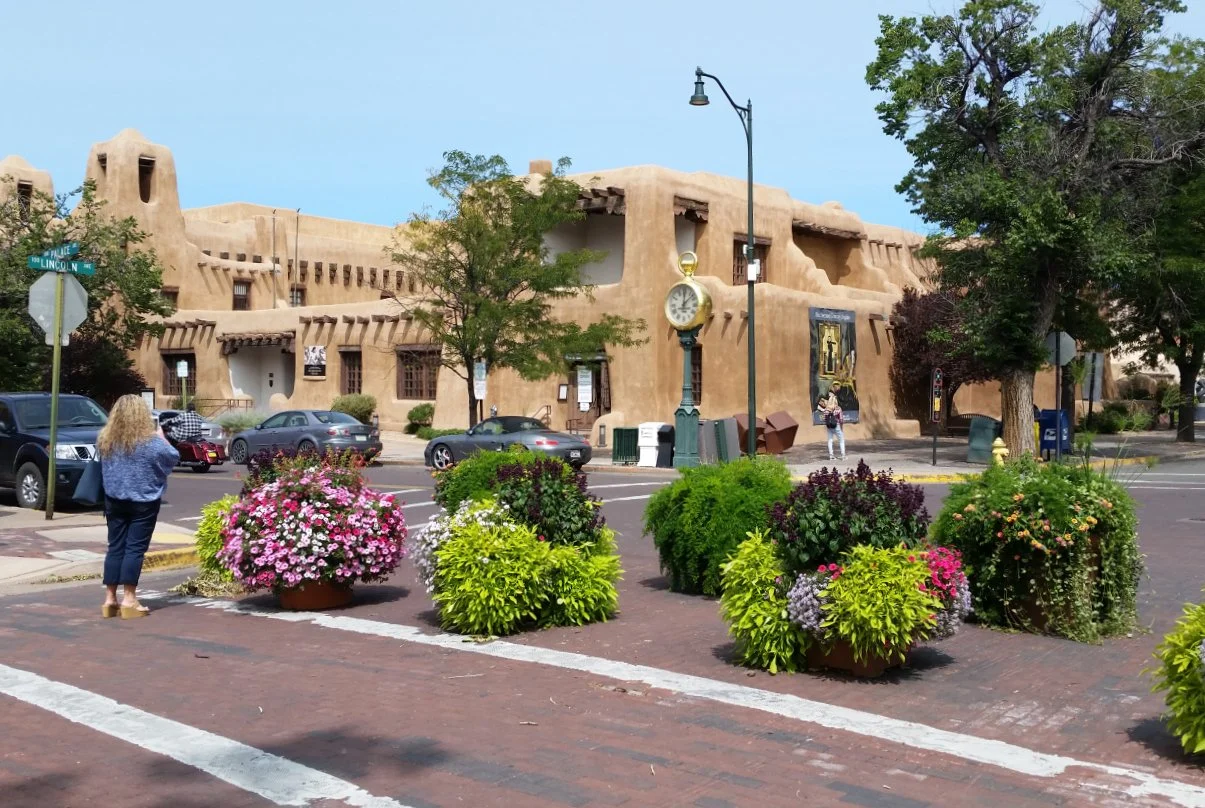 A street corner view featuring an adobe-style building, a vintage clock, colorful flowers in planters, trees, and parked cars. A woman is taking a photo or looking at her phone near the flowers, and another person is walking on the sidewalk.