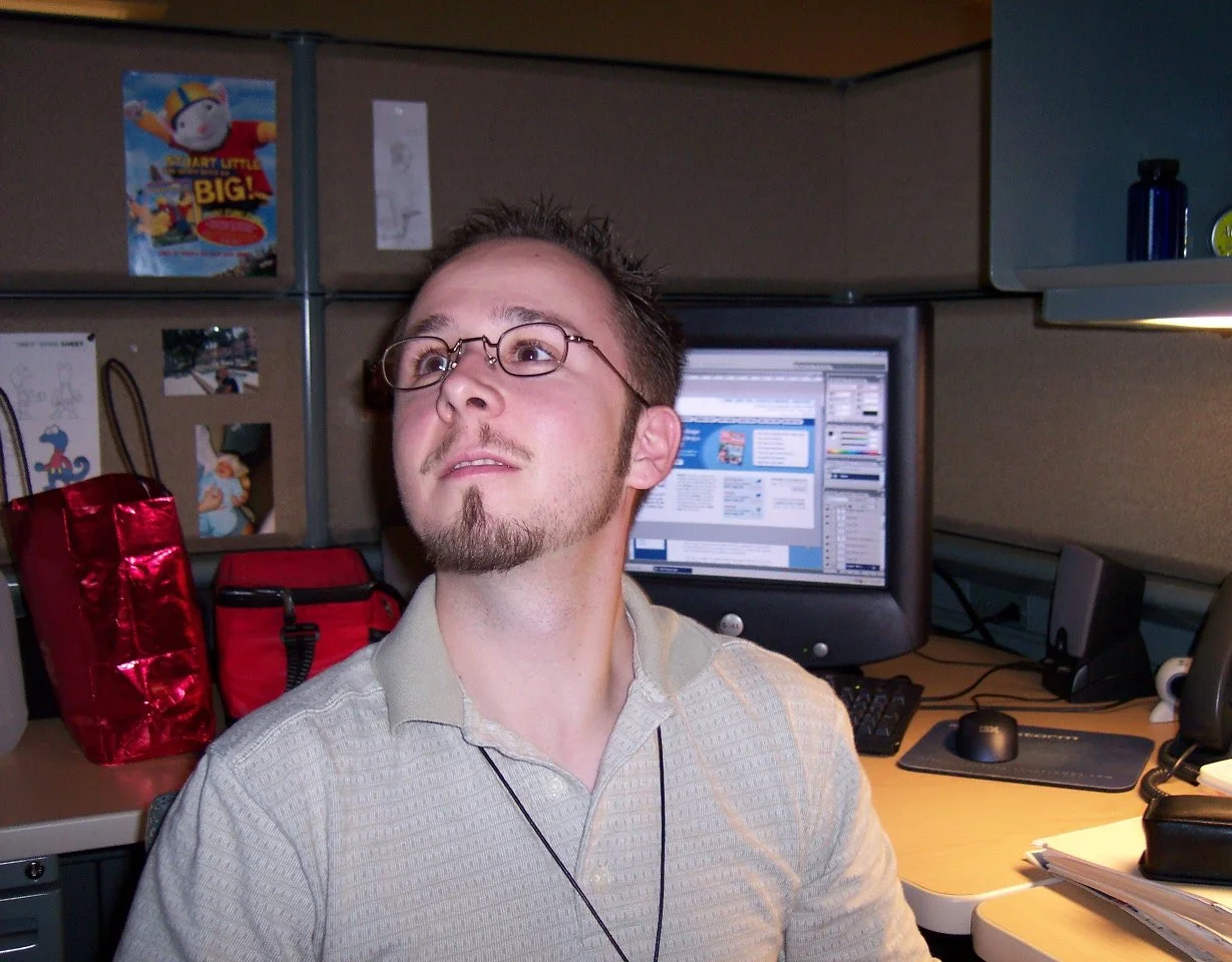 A man with glasses and a goatee sitting at an office desk with a computer and various office supplies, looking slightly upward.