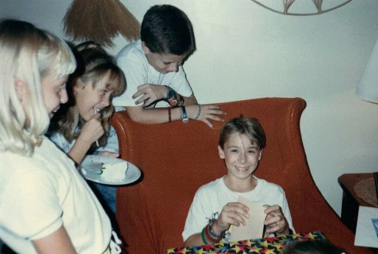 A group of children gathered around a young girl sitting on a couch, holding a birthday card and smiling at a celebration.