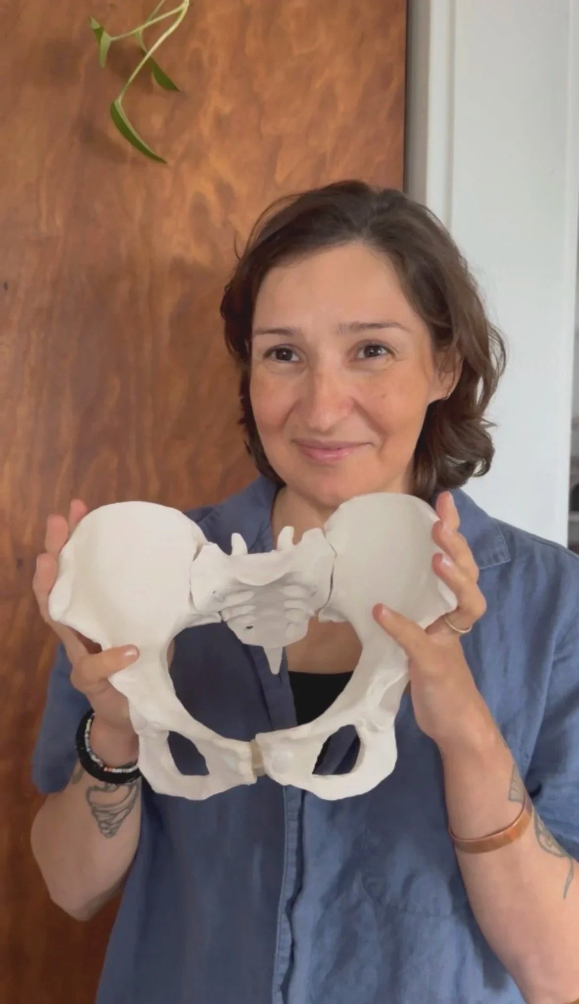 A woman with short brown hair holding a white pelvic bone model, smiling softly, standing indoors in front of a wooden wall.