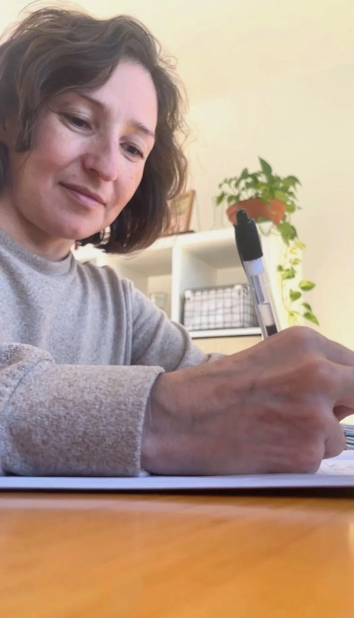 Woman sitting at a table, writing or drawing with a pen, with a plant in the background.