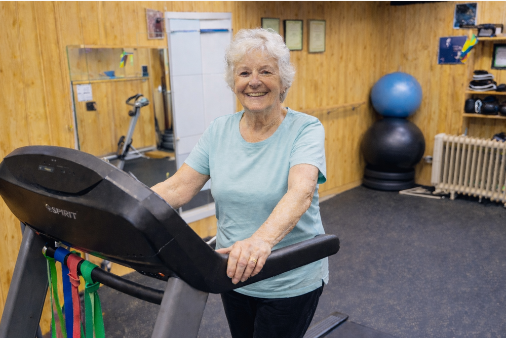 An elderly woman smiling on a treadmill in a gym with wood-paneled walls and exercise equipment.