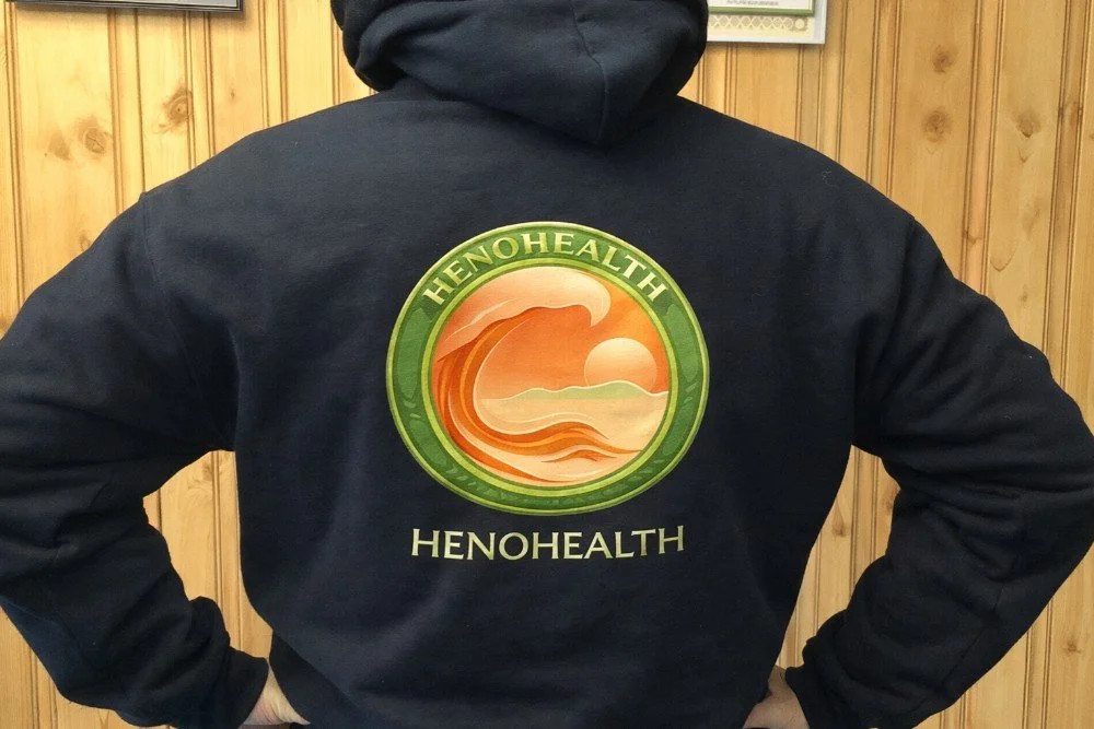 Back of person wearing a black hoodie with a large logo that reads "HENOHEALTH" and features a sunset or sunrise over water.