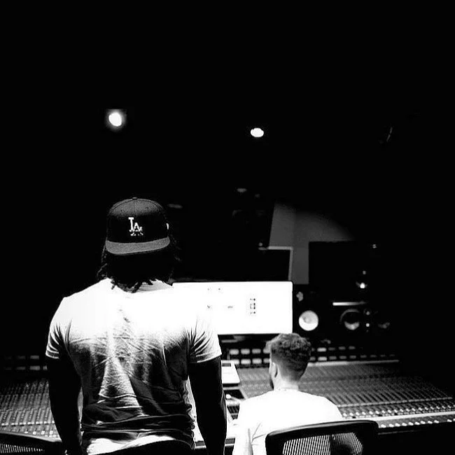 Two individuals in a recording studio, one standing with a Los Angeles Dodgers cap and the other sitting at a mixing console.