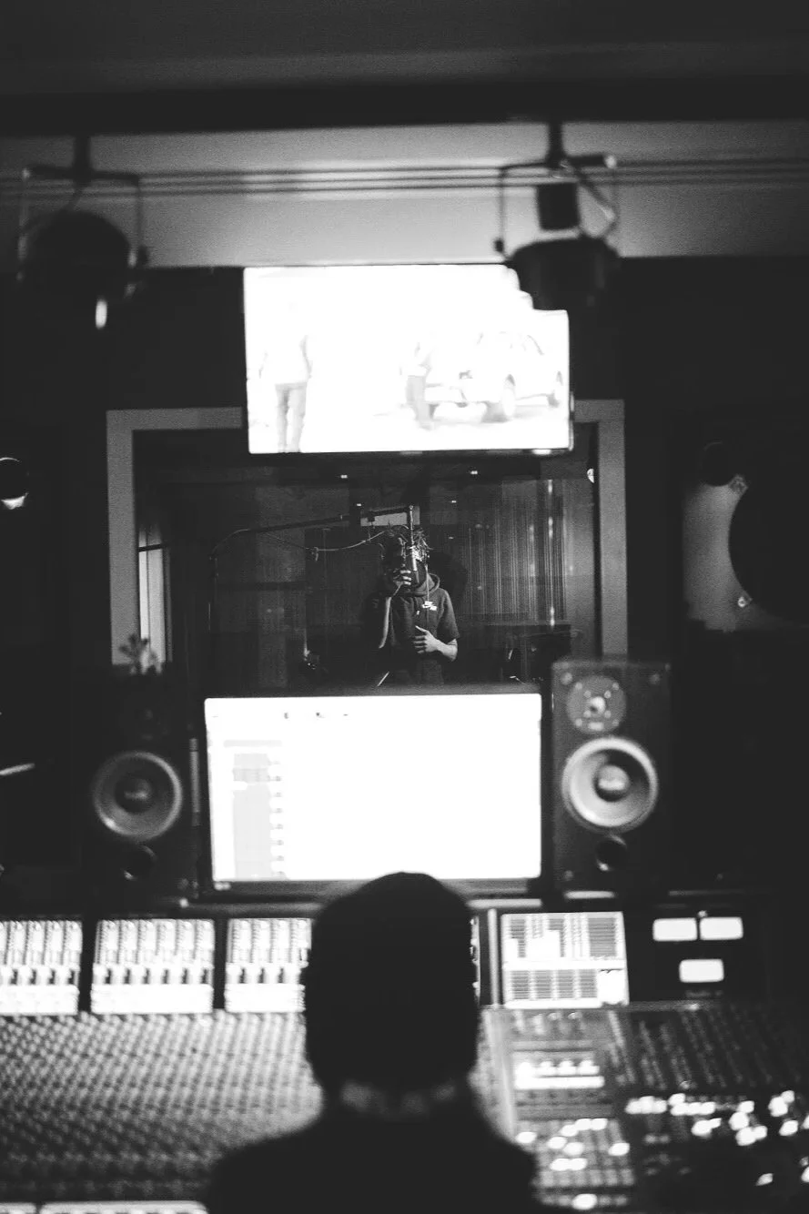 A person in a recording studio takes a selfie in front of a microphone with a pop filter. In the background, a person is visible through a glass window, and a large screen displays the person. The studio has audio monitors, mixing equipment, and a digital audio workstation interface.