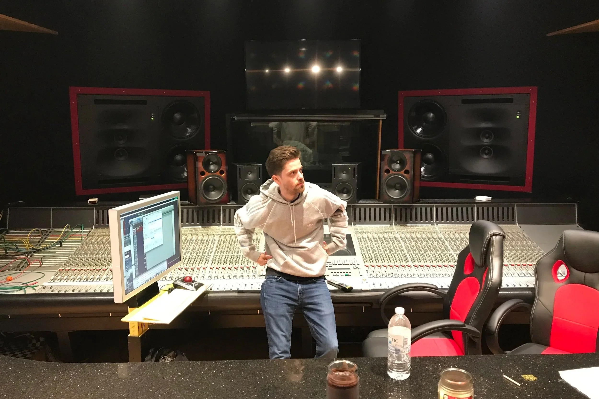 A man standing in a recording studio or sound mixing room with a large mixing console, multiple speakers, and a computer monitor on the counter in the foreground. He is wearing a light-colored hoodie and jeans.