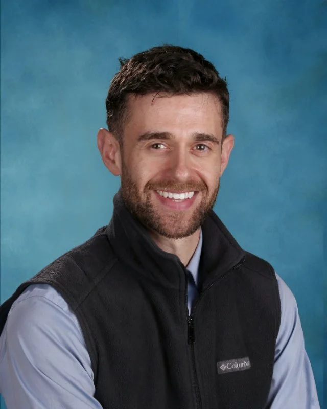 A smiling man with short dark hair, a beard, wearing a blue shirt and a black Columbia vest, posing against a blue background.