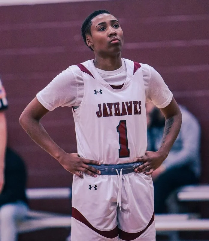 A female basketball player in a white jersey with maroon accents and the number 1, standing with hands on hips and a serious expression.