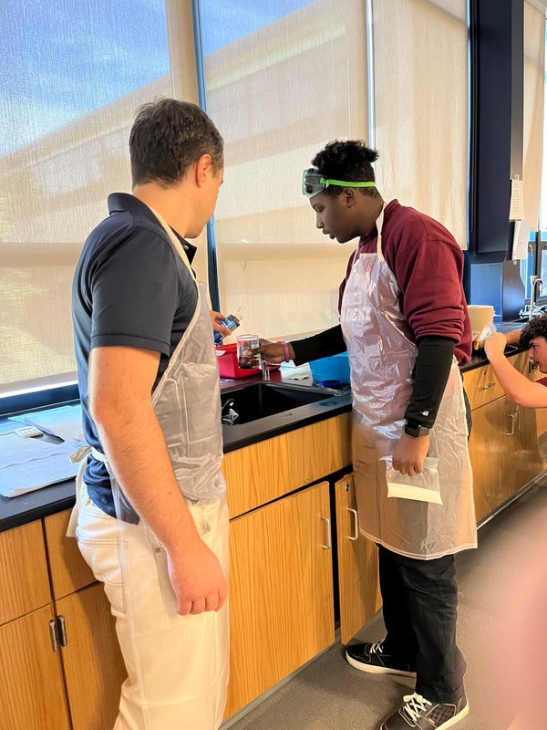 Two students wearing aprons and safety goggles conducting a science experiment at a lab sink in a classroom. One student is holding a test tube, and the other is observing.