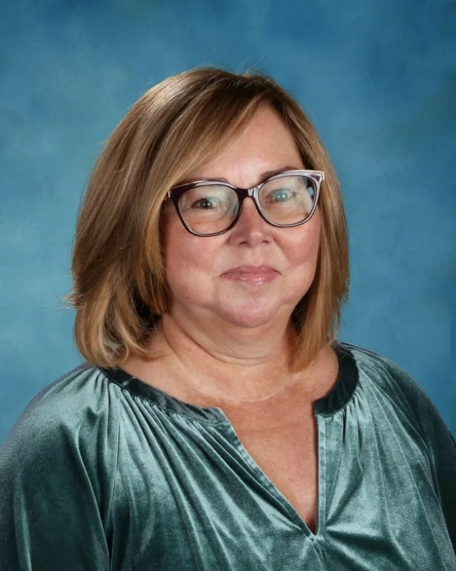 A middle-aged woman with light brown hair and glasses, wearing a shiny teal blouse against a blue background.