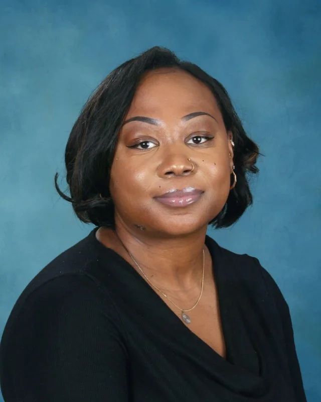 Portrait of a woman with short black hair, wearing a black top and gold jewelry, against a blue background.