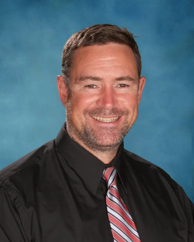 A middle-aged man with short brown hair and a beard, wearing a black judge's robe and a striped tie, smiling against a blue background.