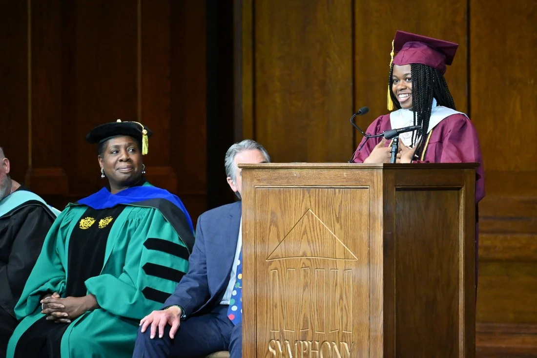 A young woman in a maroon cap and gown at a graduation ceremony, standing at a podium with a microphone, smiling, with three individuals sitting behind her, two of whom are in academic regalia.