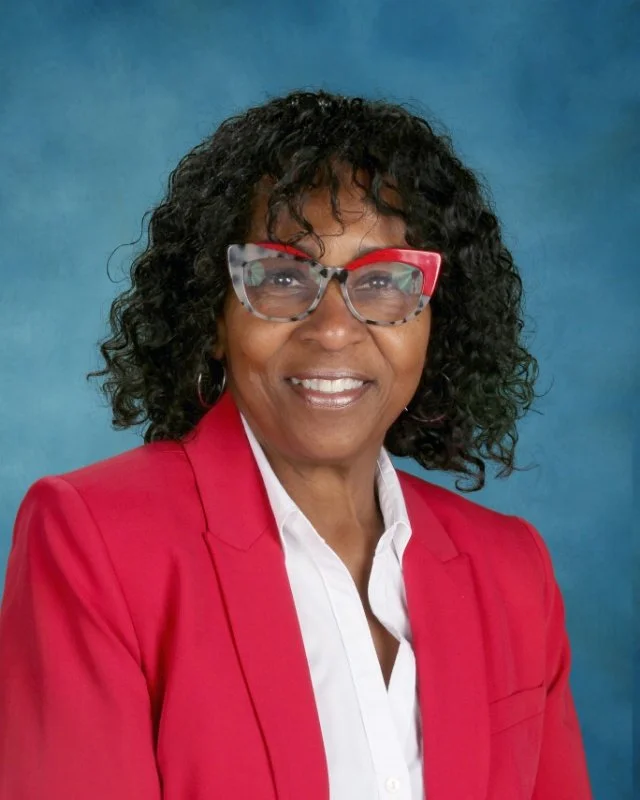 Smiling woman with curly black hair wearing red glasses, a red blazer, and a white shirt against a blue background.