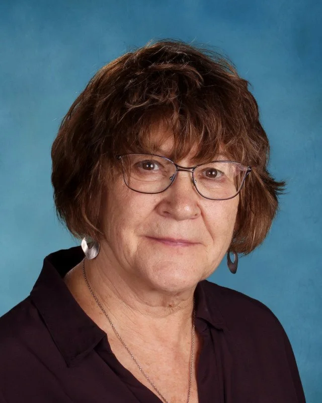 A middle-aged woman with short brown hair, wearing glasses, earrings, and a black top, posed against a blue background.