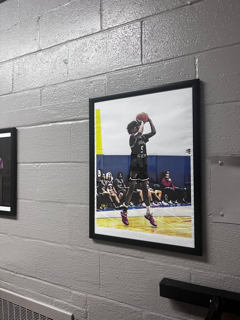 Photograph of a basketball player in mid-air taking a shot during a game, with teammates and coaches seated on the bench in the background, on an indoor court.
