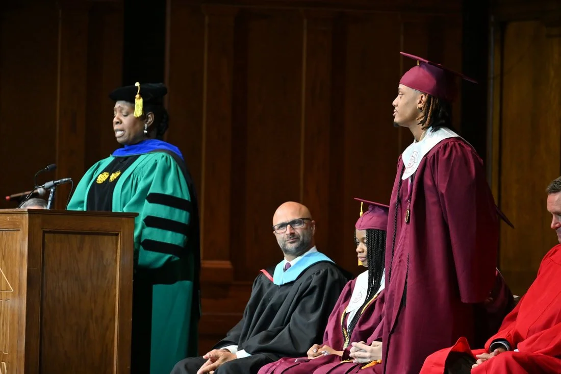 A woman in academic regalia speaking at a podium during a graduation ceremony, with four other graduates in caps and gowns seated behind her.
