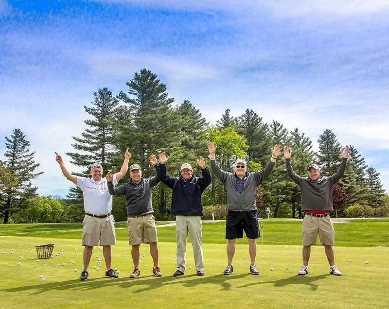 Five men standing on a golf course with their arms raised, smiling, with trees and a partly cloudy sky in the background.