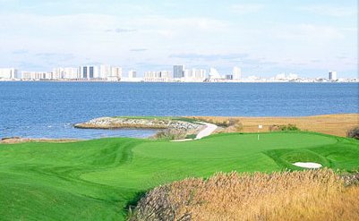 View of a coastal golf course with green fairways, a walking path, and a city skyline in the background across the water.