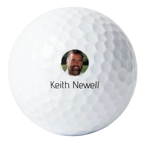 A close-up of a white golf ball with a personalized sticker that has a photo of a man with a beard and the name Keith Newell.