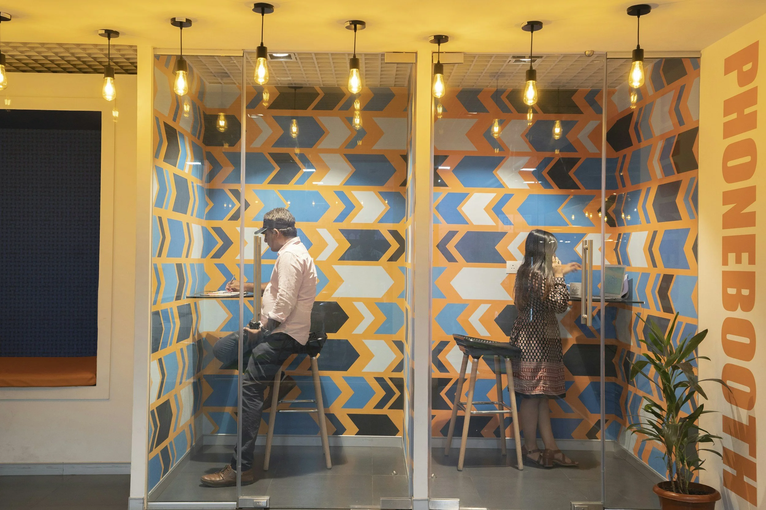 Two people sitting in separate glass booths at a photo booth shop, colorful geometric wall pattern, yellow lighting, little plant on the right.