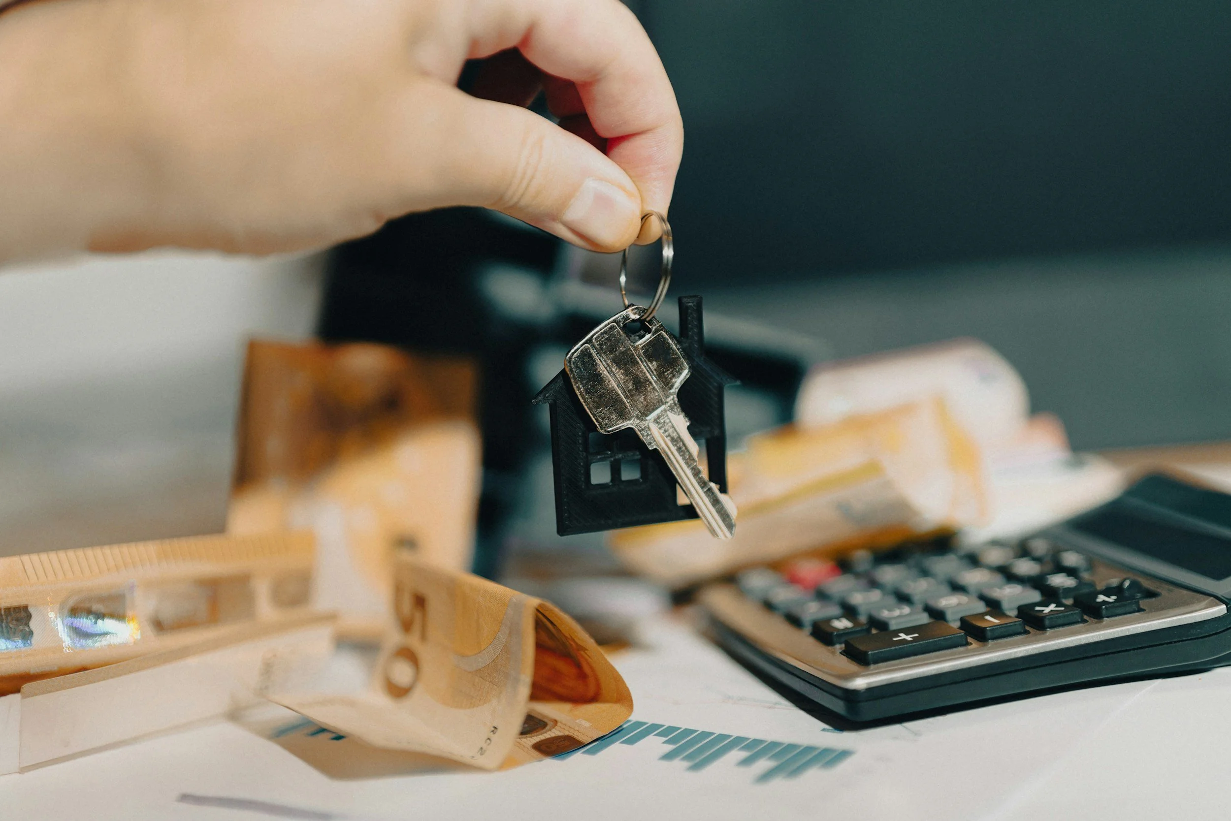 A hand holding a keyring with a house-shaped keychain and a metal key, placed near a calculator and scattered euro banknotes on a desk.