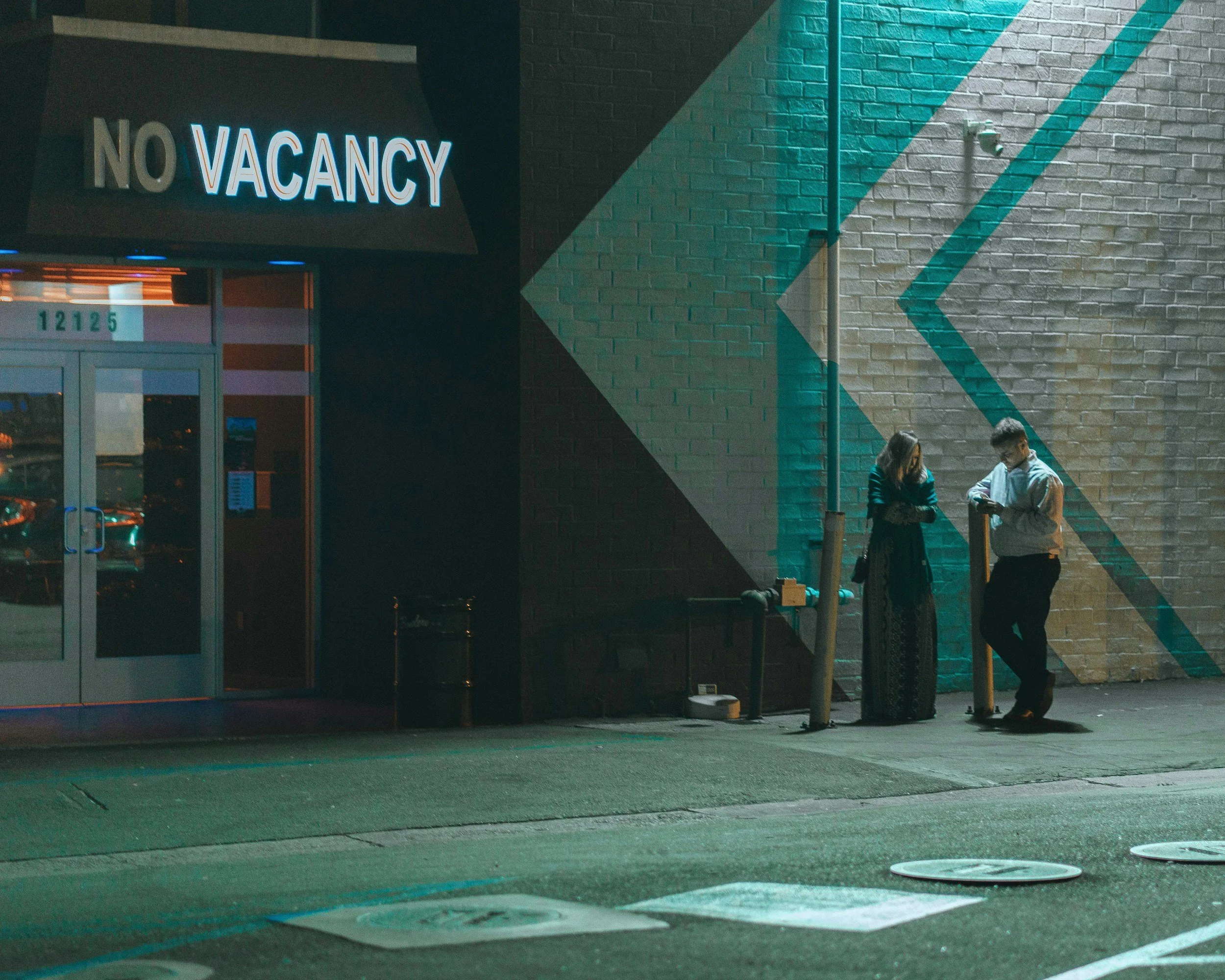 Two people standing outside a building with a neon 'No Vacancy' sign at night, having a conversation.