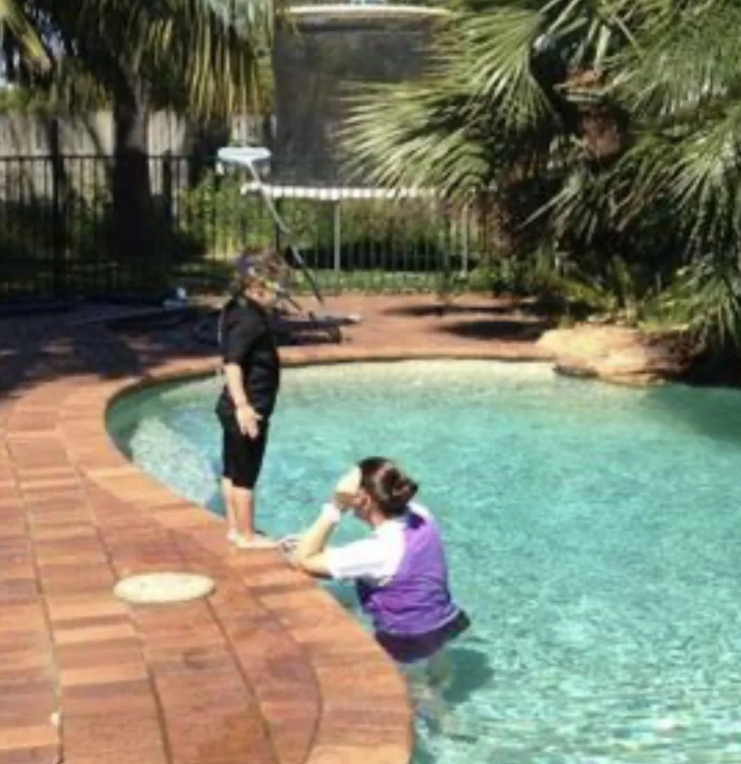 Two people by a backyard swimming pool with palm trees and a fence in the background. One person is standing on the pool deck, and the other is in the pool, resting their chin on the pool edge.
