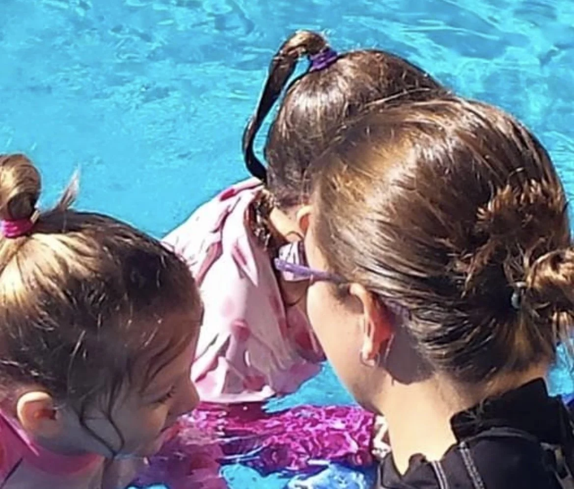 Three children with tied-back hair leaning in close to each other in a swimming pool, wearing glasses and swimsuits, with blue water in the background.