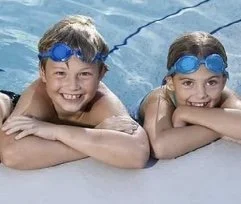 Two children wearing swimming goggles and smiling, resting their chins on the pool edge.