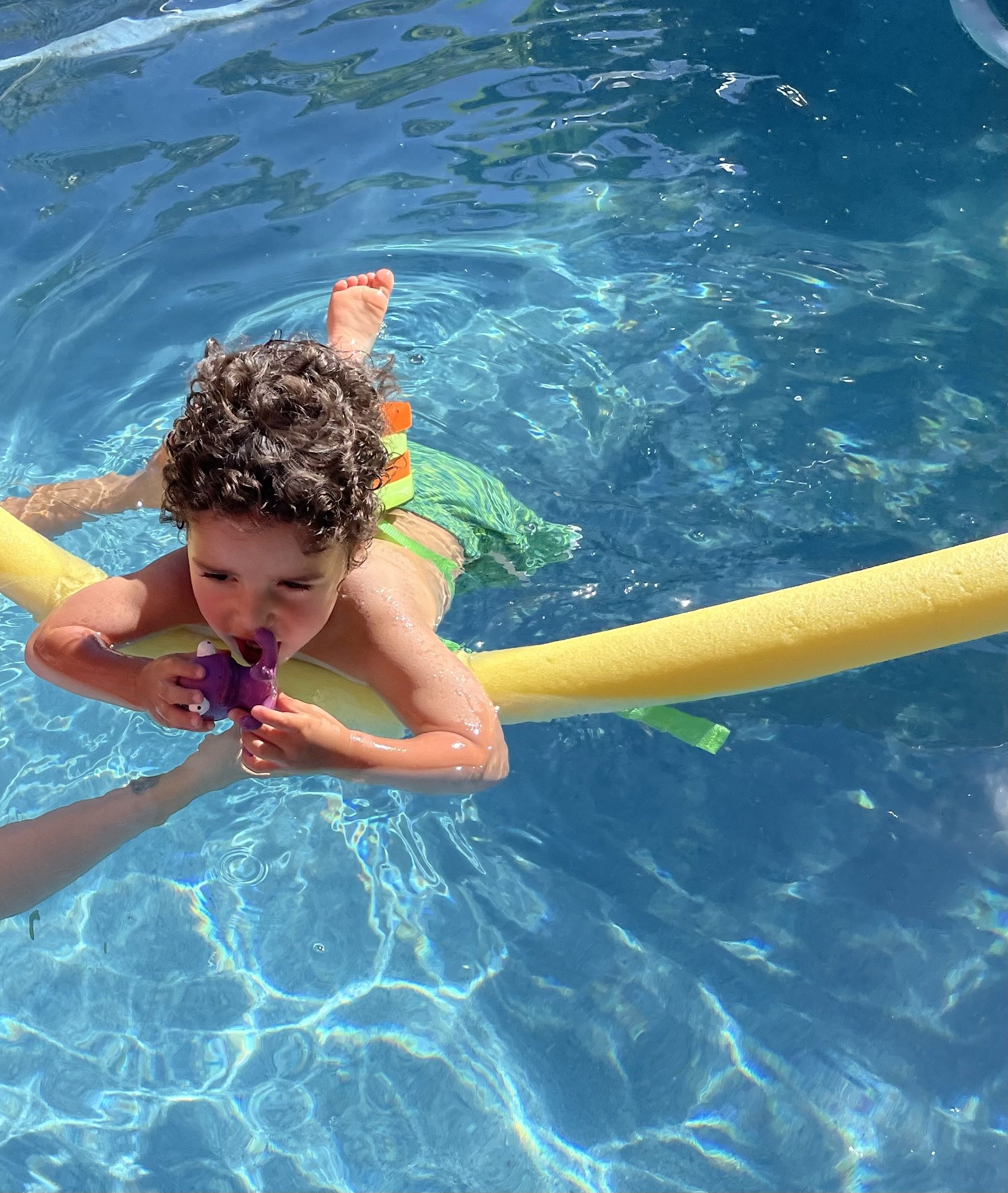 Young boy with curly hair in a swimming pool, holding a purple toy, lying on a yellow pool float.