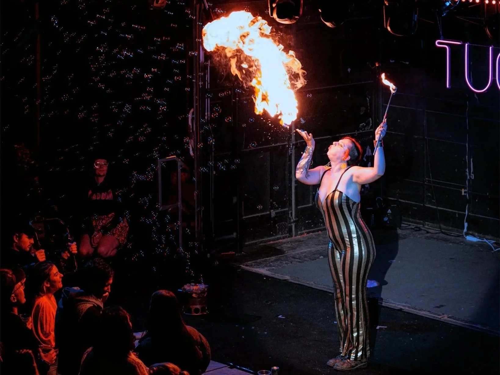 A woman in a striped, metallic jumpsuit performs a fire-breathing act on stage, with flames erupting from her mouth. The audience watches in dimly lit surroundings with a dark background and neon pink letters partially visible.