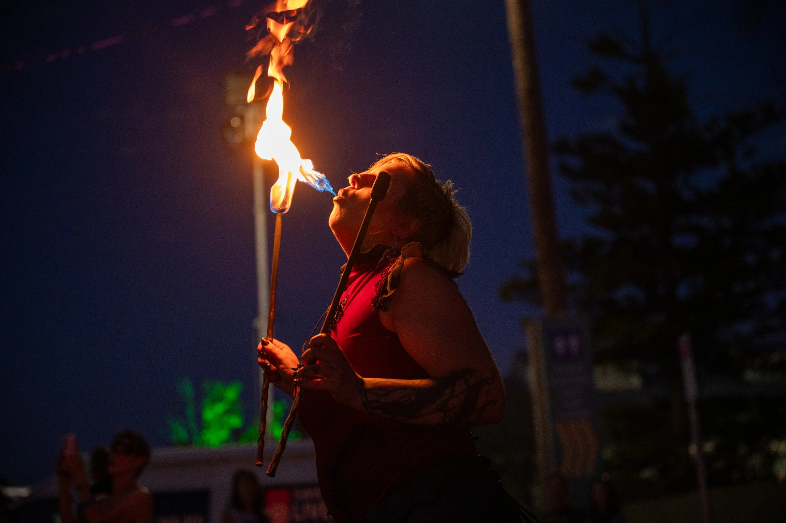 A woman with short hair and tattoos on her arms is performing a fire-eating act at night, holding a lit torch near her mouth, creating flames.