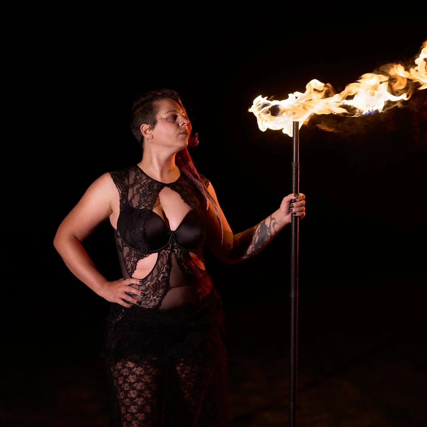 A woman in black lace clothing holds a flaming torch in her right hand, standing against a dark background.