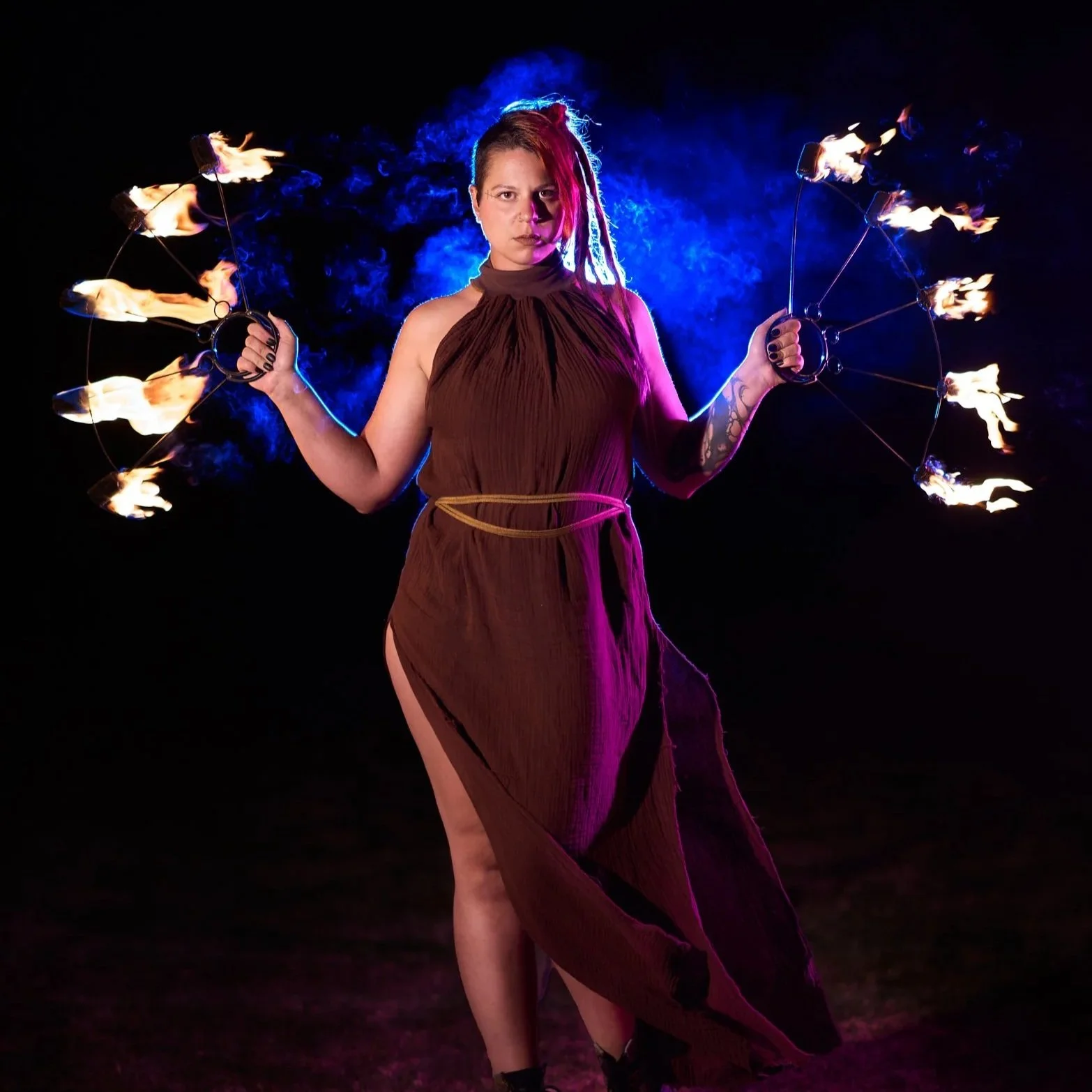 A woman with dreadlocks holding fire fans creating a fiery display in the dark, with blue smoke behind her.