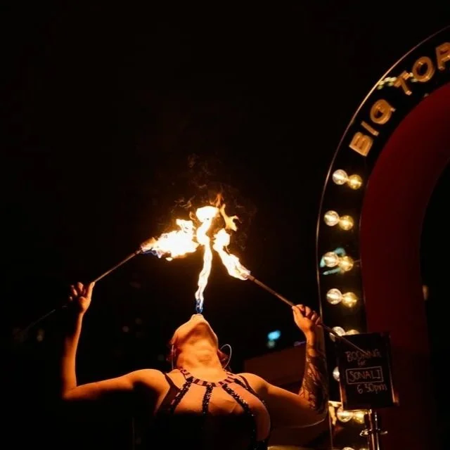 A female fire performer in a black and pink costume spins flaming batons at night, with a sign for Big Top  and a small chalkboard indicating a show time of 6:30 pm.