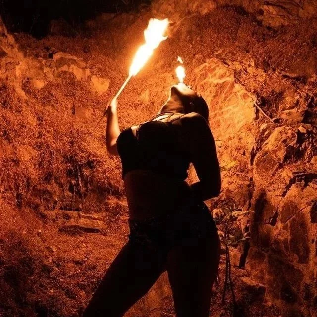 A person performing a fire breathing act at night, with flames coming out of their mouth and a torch in one hand, against a rocky background.